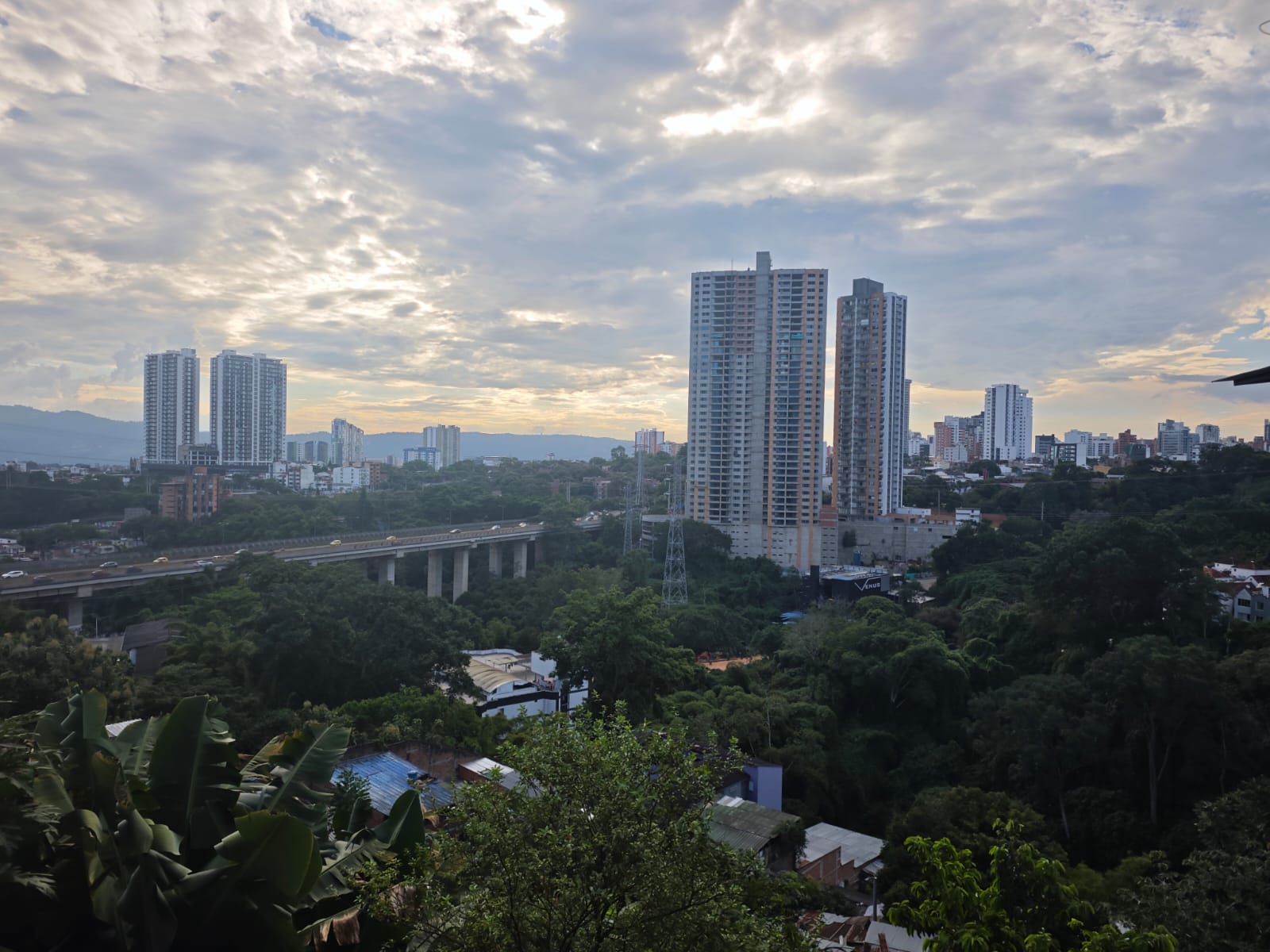Vista panorámica de Bucaramanga al atardecer, con edificios de gran altura, el viaducto de acceso a la ciudad y zona verde en primer plano